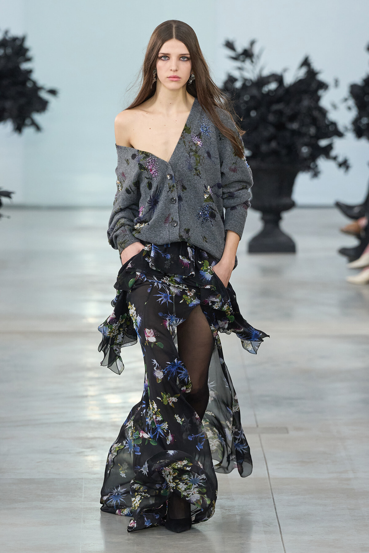 Model on a fashion runway wearing an off-shoulder gray cardigan with floral embroidery and a sheer black floral-printed skirt.