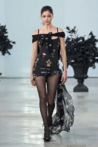 Model walking runway in a black floral off-shoulder mini dress with sheer train and tights.