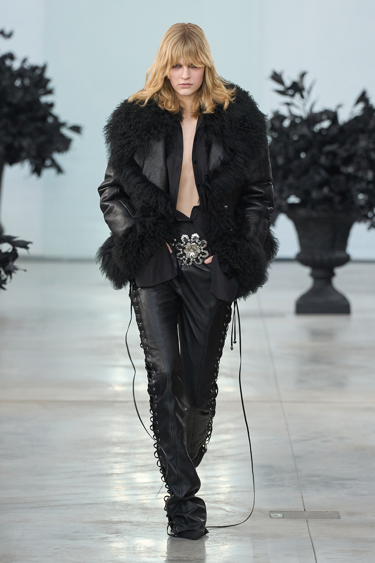 Model walks the runway in a black shearling coat with voluminous fur, open at front, paired with black lace-up leather pants and a jeweled belt centerpiece.