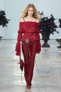 Model walks a fashion runway in an off-shoulder red blouse with ruffles and matching velvet red pants, accessorized with a silver floral belt.