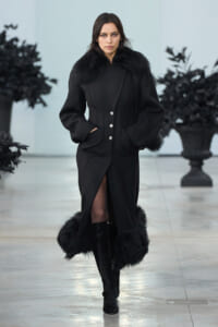 Model walking a runway in a long black coat with fur trim at the collar and cuffs, hands in pockets