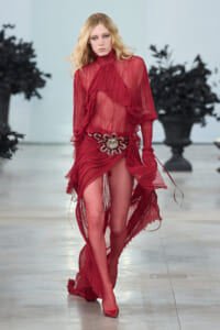 Model walking a runway in a sheer red ruffled dress with a large jeweled belt.