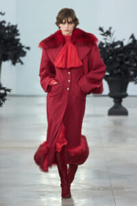 Model on a runway wearing a crimson fur-trim coat with large lapels, red scarf bow, and tall red boots.