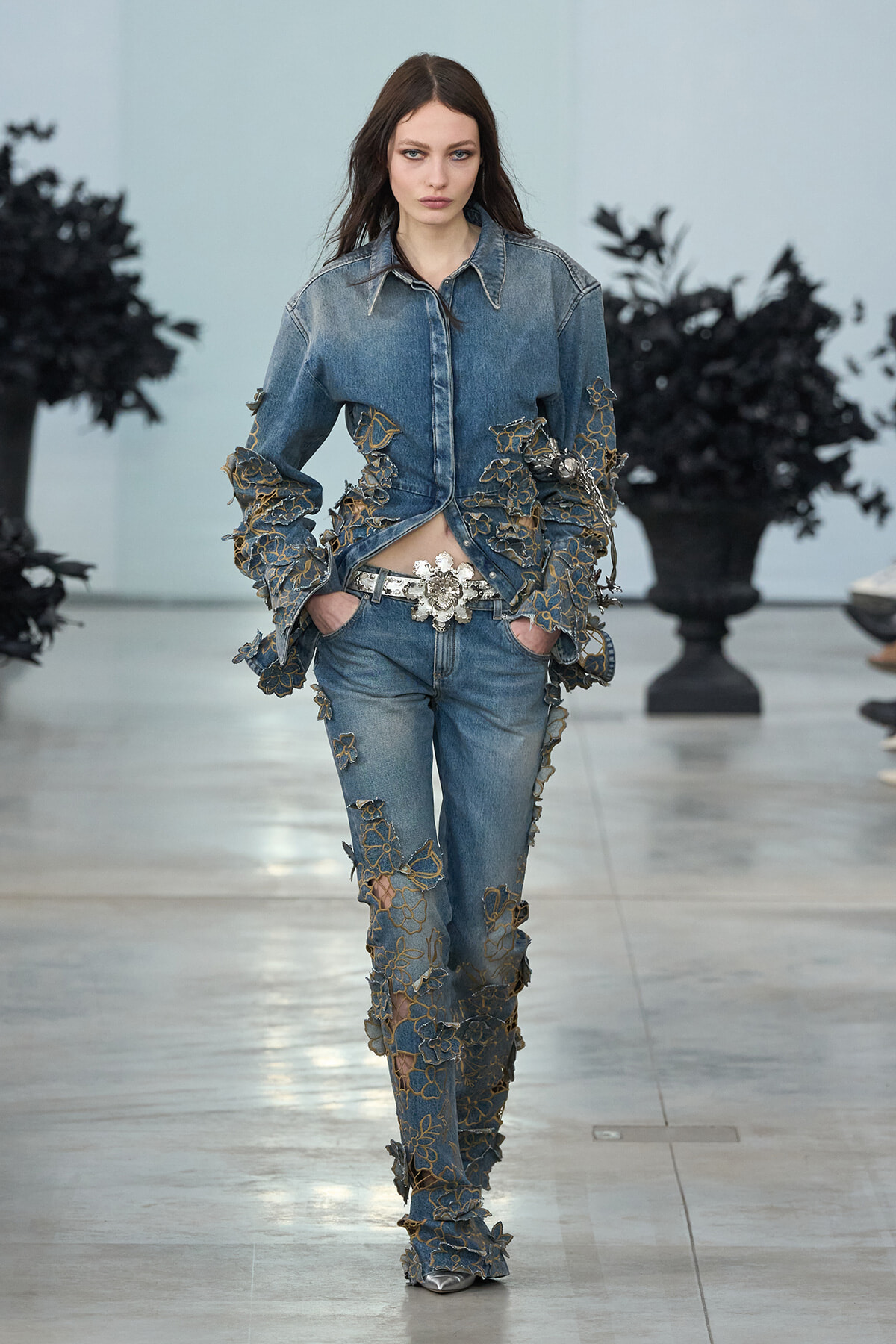 Model walking the runway in a denim-on-denim ensemble with gold floral lace appliqués from jacket to boots, silver belt, and metallic shoes.