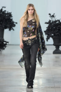 Model walks the runway in a sheer floral one-shoulder top and dark jeans with a large decorative belt.