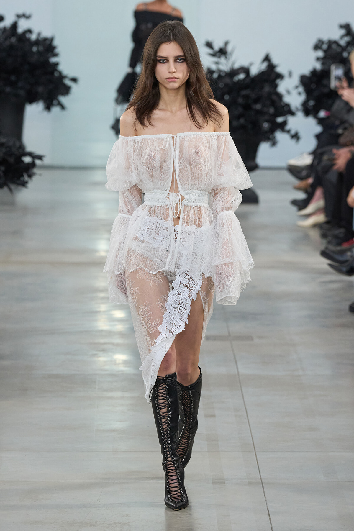 Model walking a runway in a white off-shoulder lace dress with a tied waist, sheer layers, and black lace-up knee-high boots.