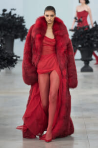 Model walking the runway in a voluminous red fur coat over a sheer red dress.