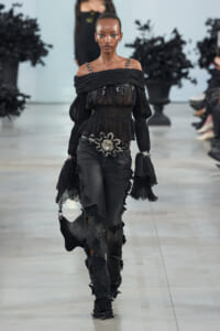 Model walking a runway in a sheer black off-shoulder blouse with ruffled sleeves, wide belt, and dark floral-embellished pants with matching accessories