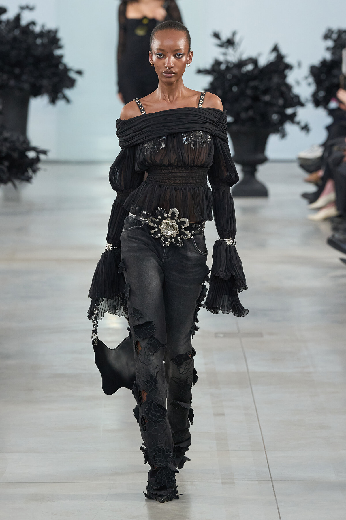 Model walking a runway in a sheer black off-shoulder blouse with ruffled sleeves, wide belt, and dark floral-embellished pants with matching accessories