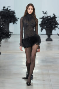 Model walks a runway in a black sheer lace turtleneck top with a fluffy black fur mini skirt and sheer tights with embellished heels.