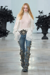 Model walking the runway in a sheer white ruffled blouse, ornate belt, and jeans with gold floral embroidery along the legs.