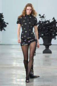 Female model walks a runway in a black floral short-sleeve top with a structured peplum and matching skirt with sheer floral wrap, black tights, and jeweled heels.