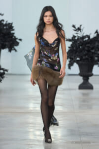 Model walks the runway in a sheer black floral slip dress with a brown fur mini-skirt and black tights.