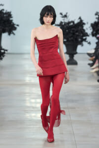 Model walking a runway in a red strapless, structured top with matching red tights and red shoes.