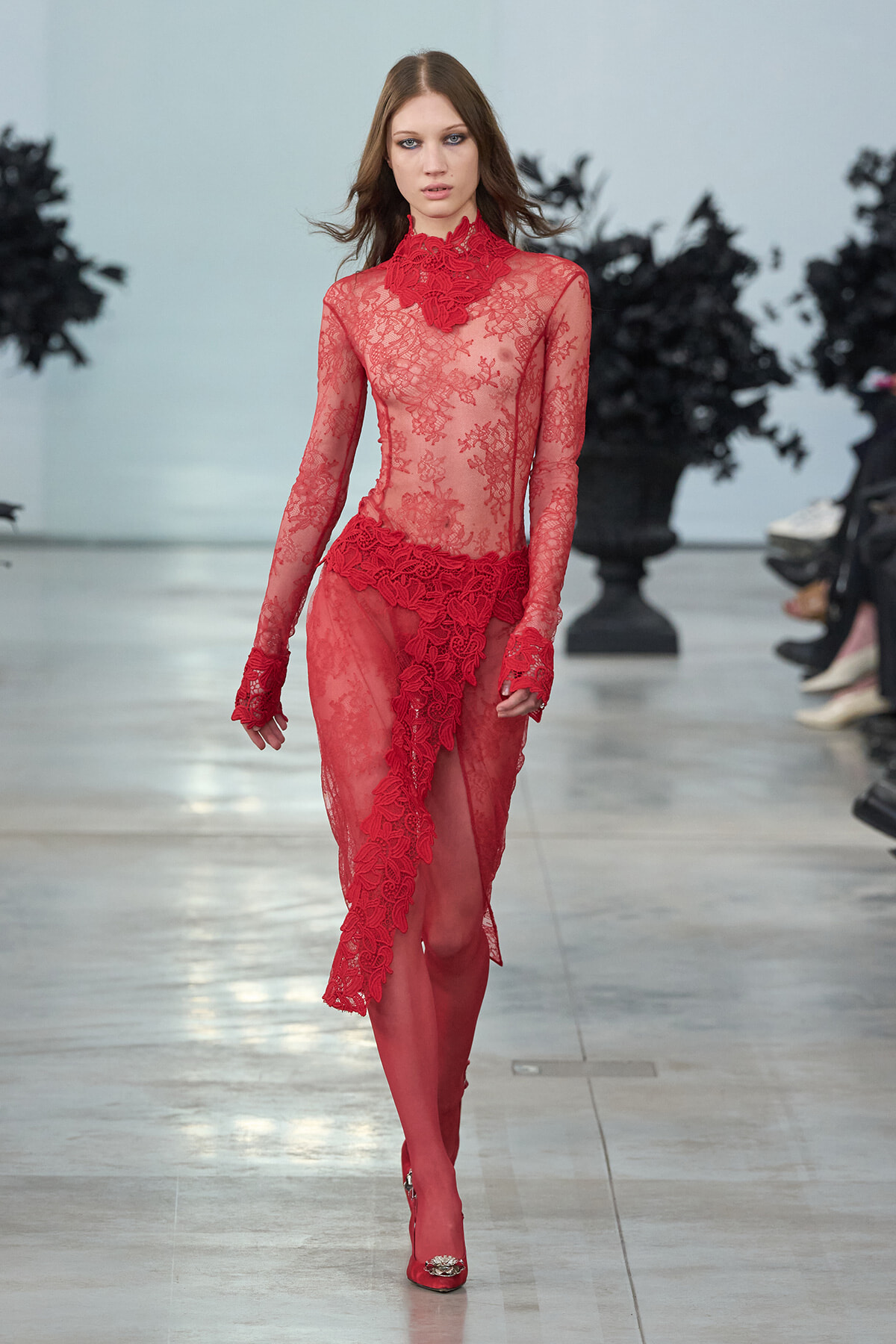 Model walking a runway in a sheer red lace gown with high neckline and floral embroidery, matching gloves, and red heels.