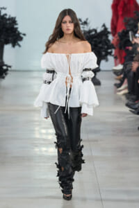 Model walking a runway in a white off-shoulder blouse with tie fronts and black floral-embellished leather pants, on a fashion show catwalk.