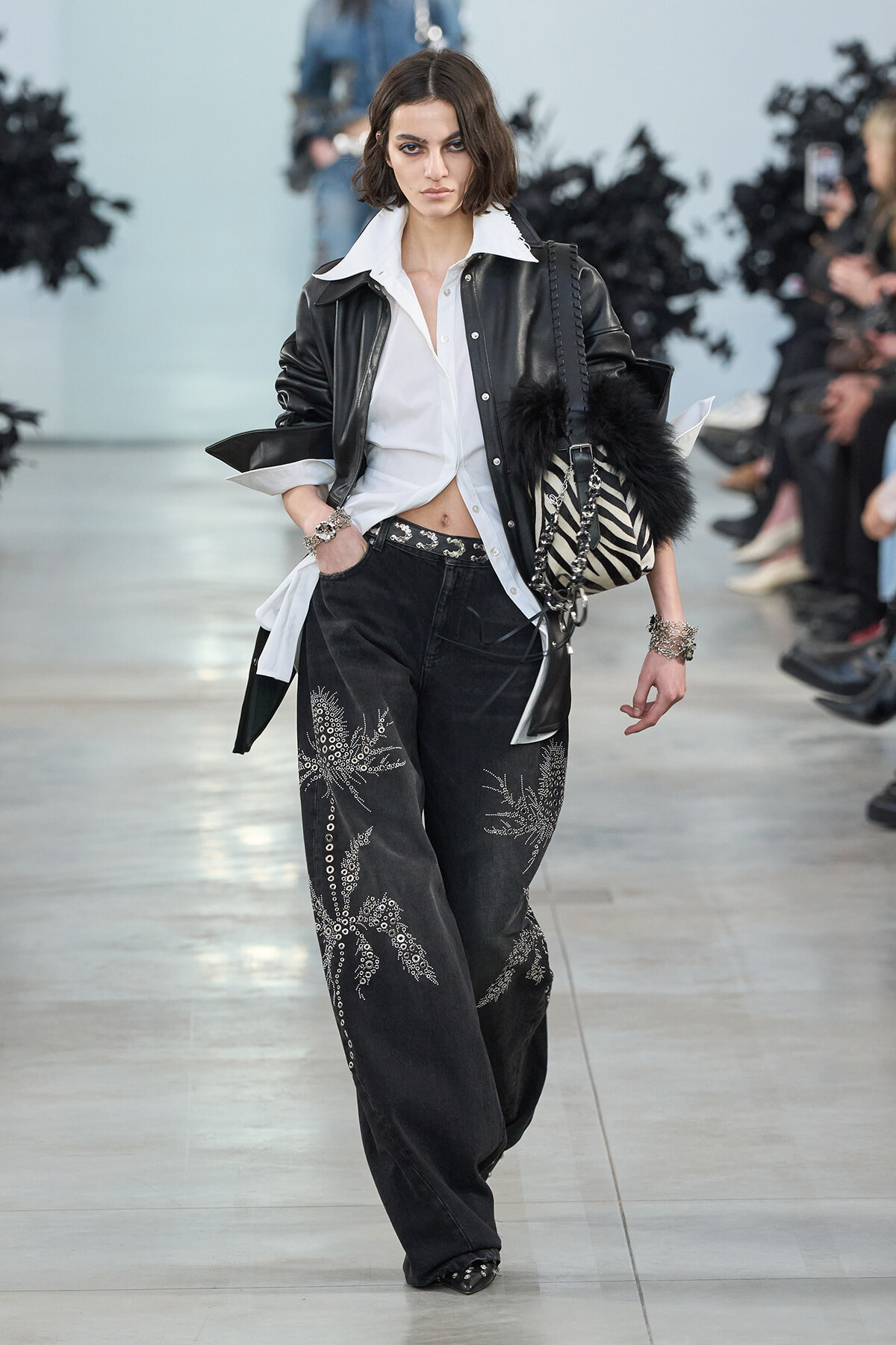 Female model on a runway wearing a white shirt tucked into embroidered black wide-leg jeans, a black leather jacket, and a zebra-striped bag with fur detail.