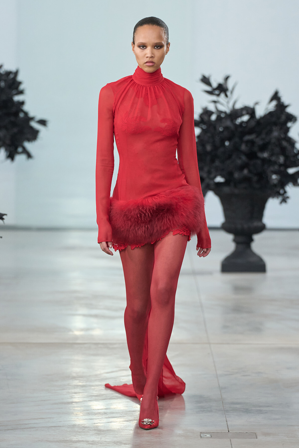 Model walks the runway in a red sheer turtleneck dress with red fur trim at the hem.