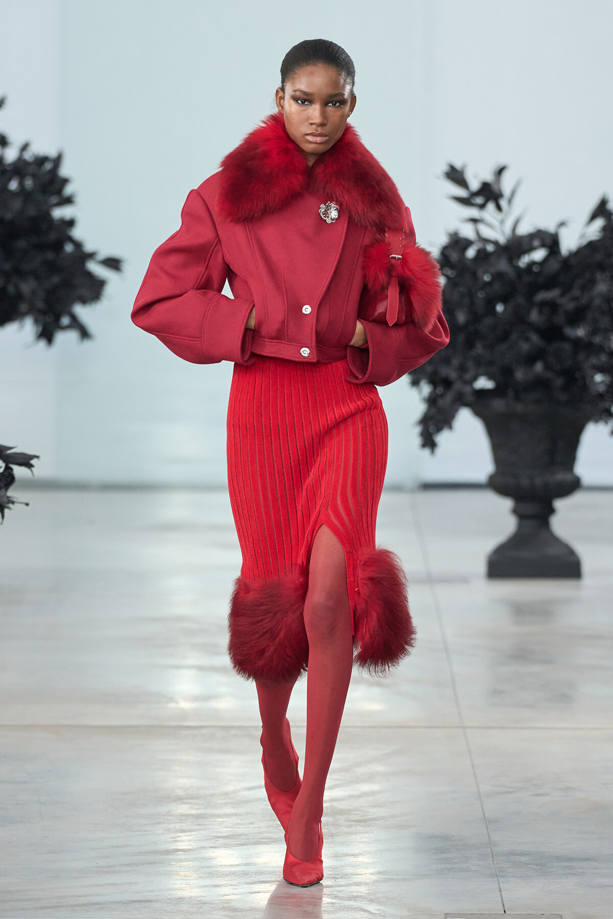 Model walking a runway in a striking red fur-collared coat and matching ribbed red skirt with fur accents at the hem and cuffs.