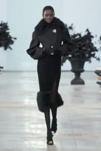 Model walking a runway in a black fur-collared coat with a brooch, a pleated velvet midi skirt with a front slit, and fur-trimmed cuffs with embellished heels.