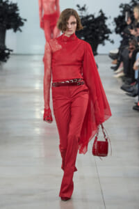 Model walking runway in red lace-shouldered top with cape, matching red trousers, black belt, and red handbag.