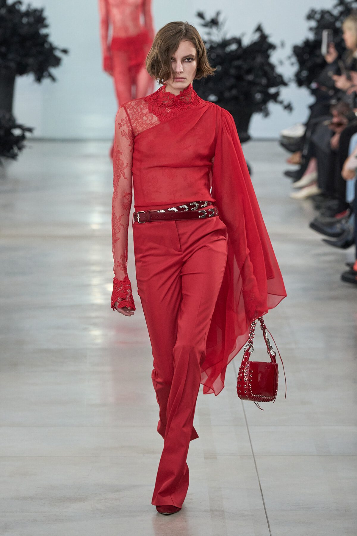 Model walking runway in red lace-shouldered top with cape, matching red trousers, black belt, and red handbag.