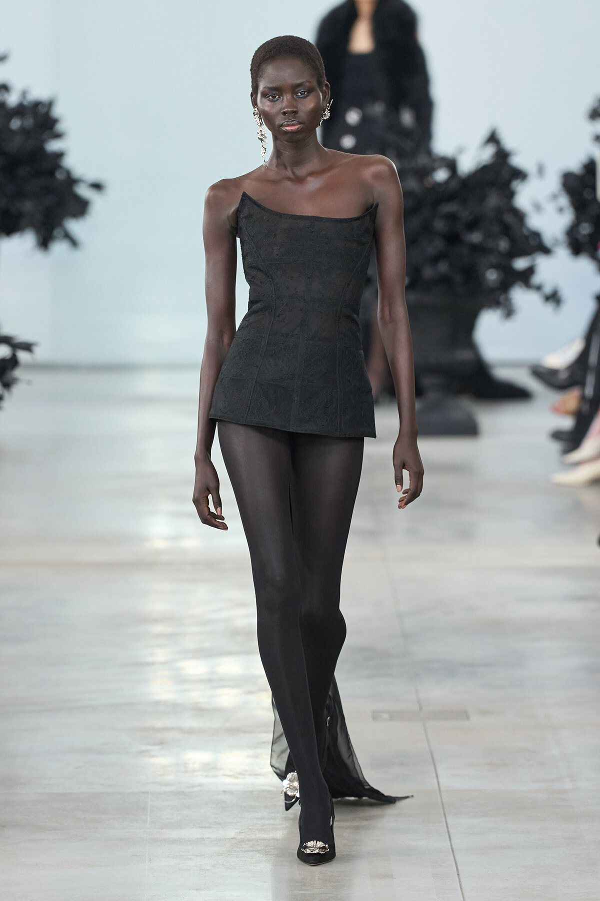 Model walks the runway in a black strapless mini dress paired with opaque black tights and embellished heels, wearing statement earrings.
