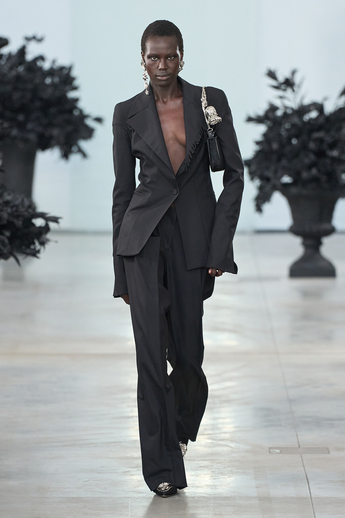 Model wearing an all-black tailored pantsuit with a deep V-neck and oversized blazer, walking a runway with a black handbag and statement earrings.
