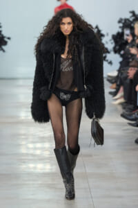 Model on a runway wearing a voluminous black faux-fur coat, sheer top, black underwear with a butterfly design, sheer tights, and studded boots, carrying a studded handbag.