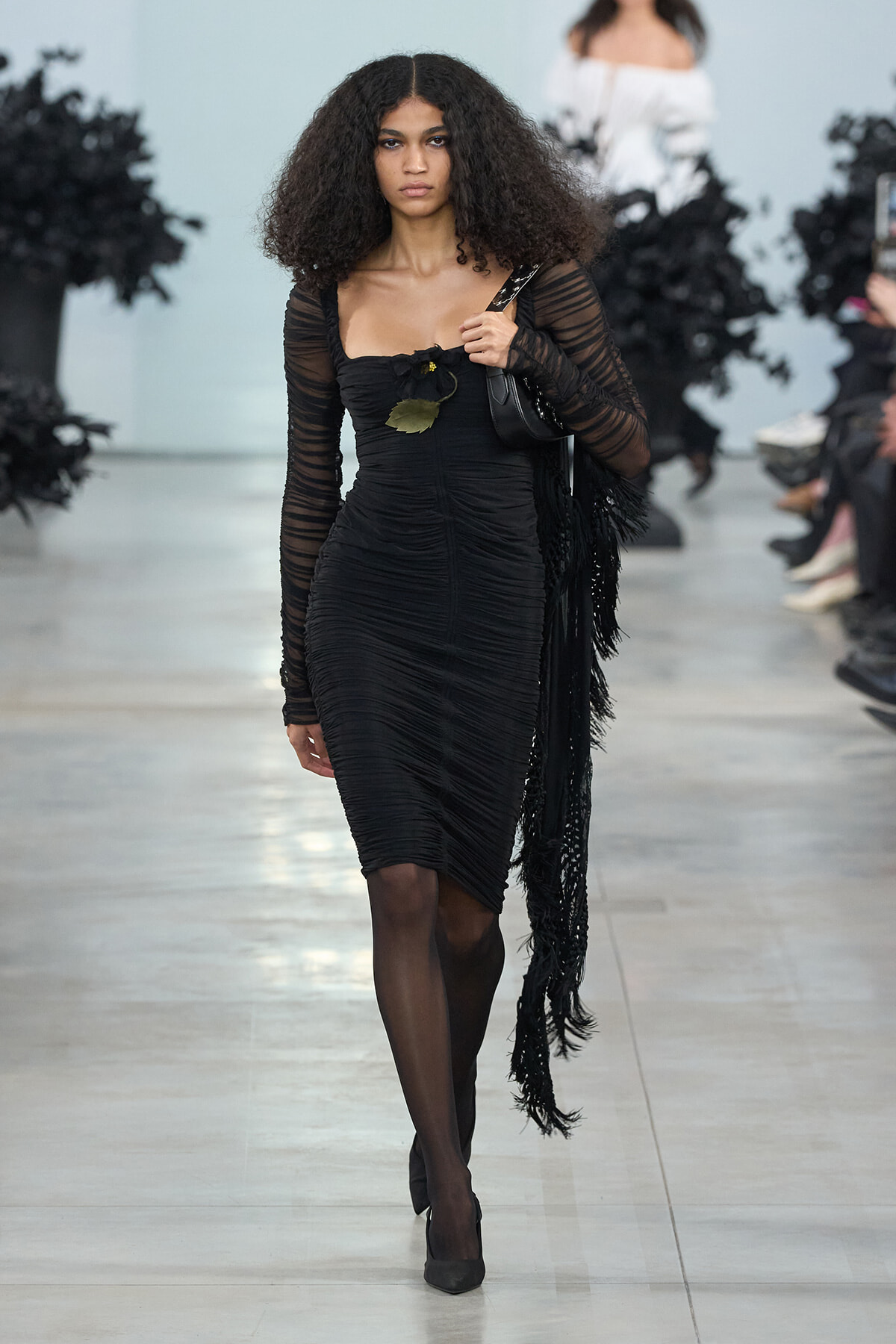 Model walking a runway in a black ruched midi dress with sheer long sleeves, carrying a black shoulder bag and a fringed scarf.