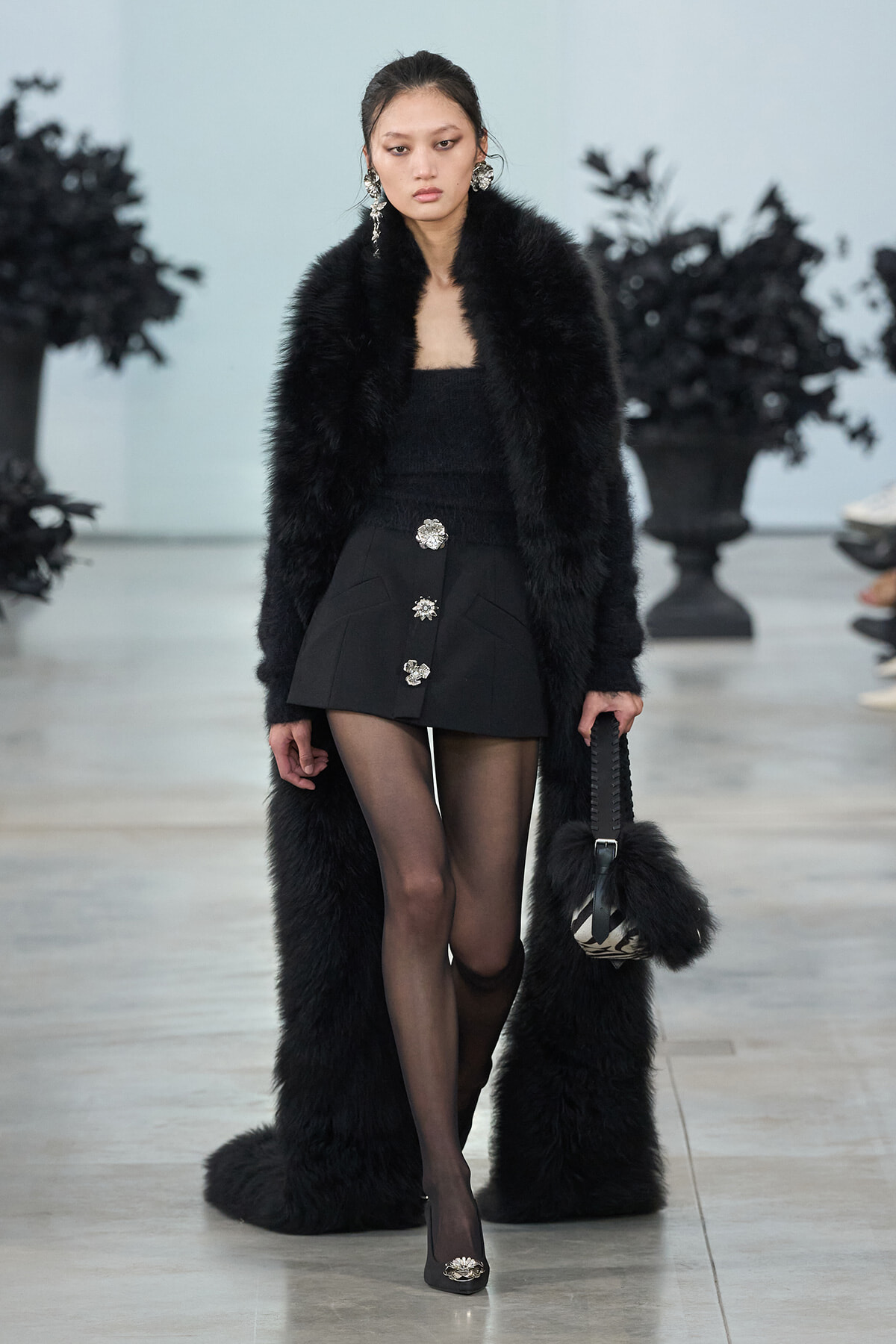 Model walking a runway in a long black fur coat over a black outfit with three silver brooches, carrying a furry zebra-print bag.