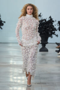 Model walks a fashion show wearing a sheer pale pink gown densely covered in white floral appliqués, with long sleeves and a high neck, paired with silver heels on a runway.
