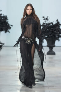 Model walking a runway in a sheer black ruffled gown with a high neck and ornate belt, black boots visible beneath the flowing fabric.