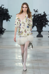 Model walks a fashion show wearing an off-shoulder cream floral cardigan and a white fluffy mini skirt, with silver heels and a metallic handbag.