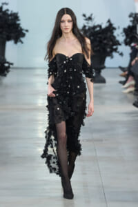 Model walking the runway in a black off-shoulder gown with sheer overlay and 3D floral embellishments.