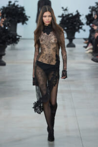 Model walking a runway in a sheer black lace gown with long sleeves and floral embroidery on the dress, high-neck design.