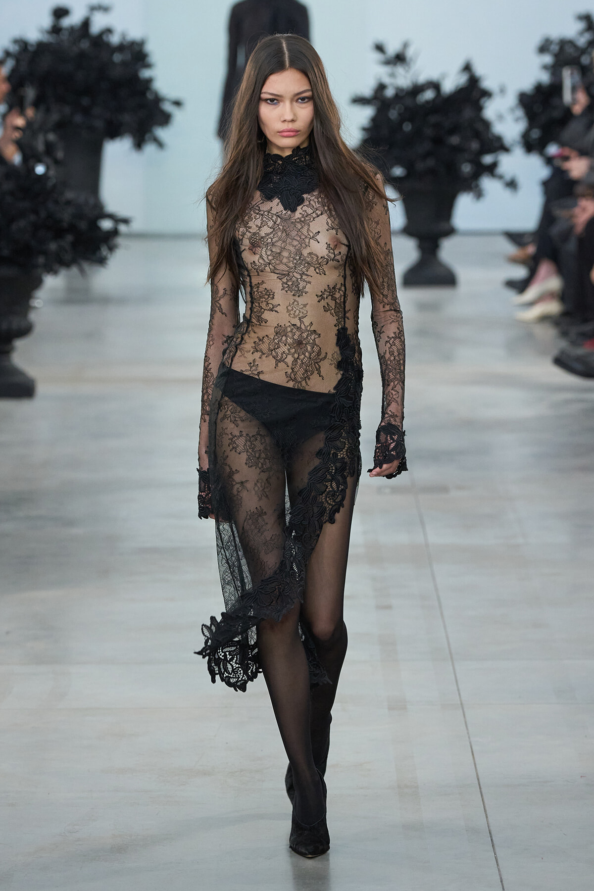 Model walking a runway in a sheer black lace gown with long sleeves and floral embroidery on the dress, high-neck design.