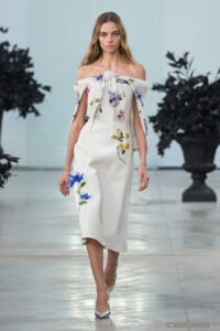 Model walks the runway in a white off-the-shoulder midi dress with blue and yellow floral embroidery and a tied floral cape draped over her arms, silver heels.