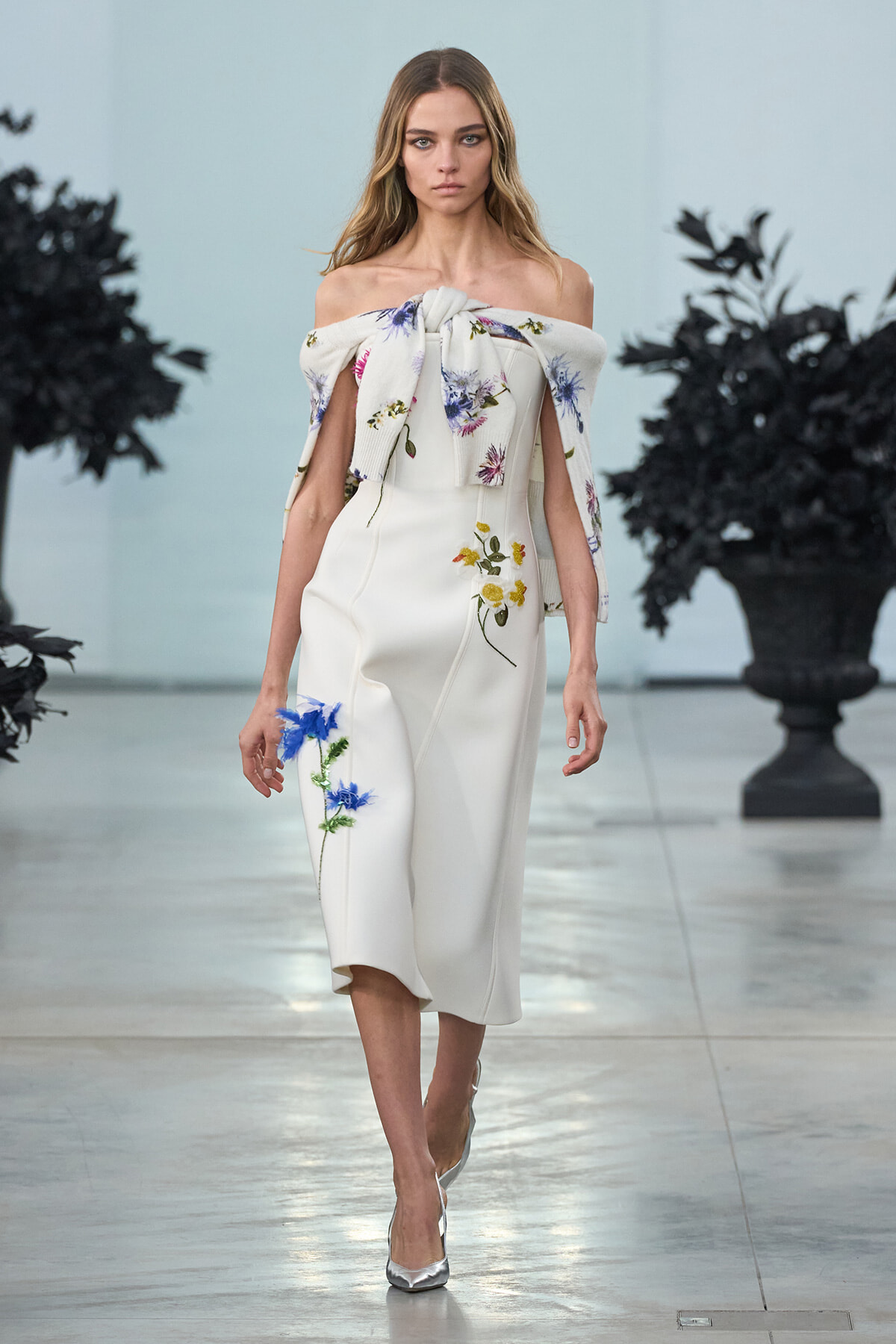Model walks the runway in a white off-the-shoulder midi dress with blue and yellow floral embroidery and a tied floral cape draped over her arms, silver heels.