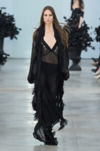 Model walking the runway in a sheer black gown with lace bodice and long fringe-trimmed outerwear.