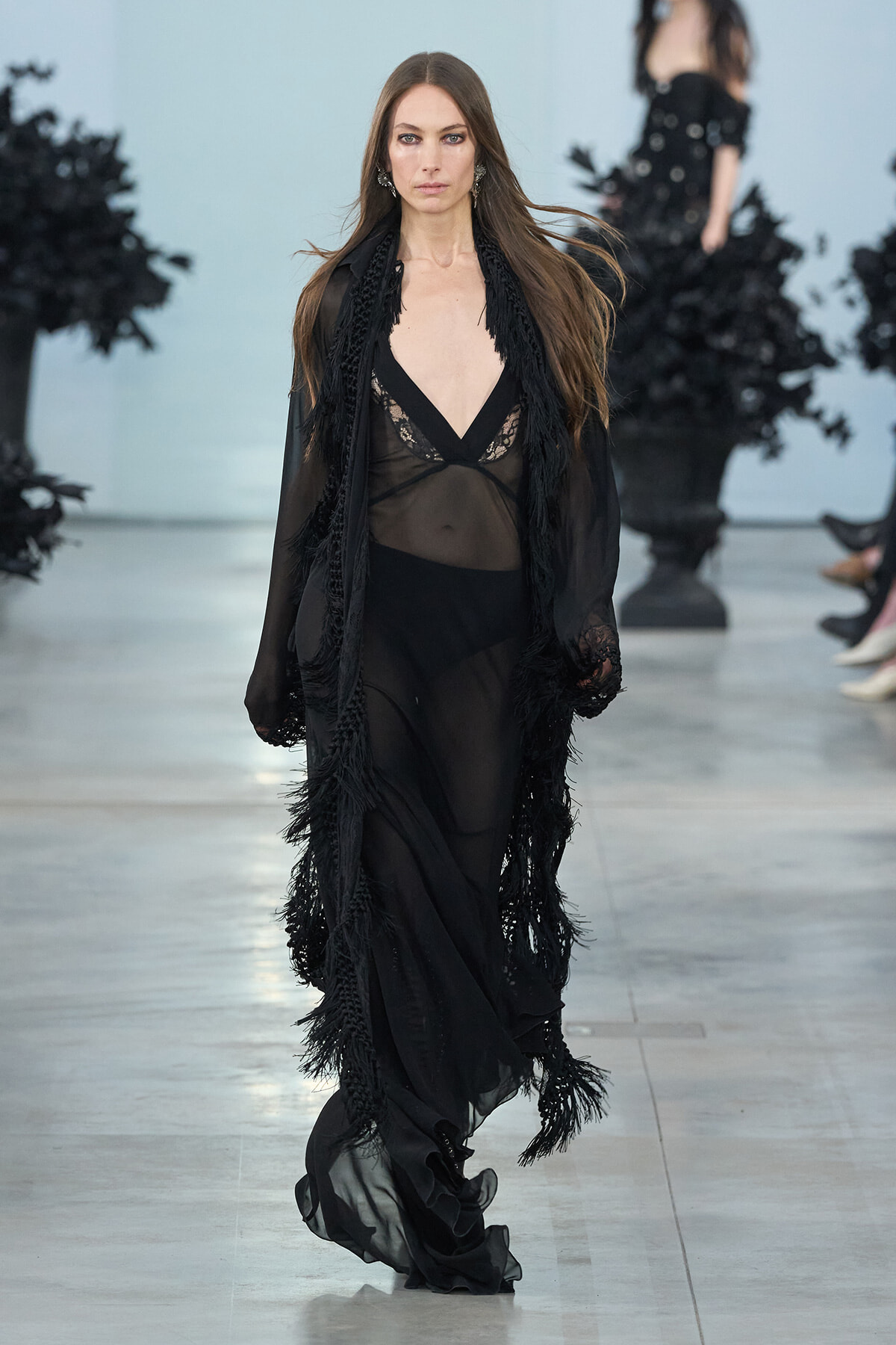 Model walking the runway in a sheer black gown with lace bodice and long fringe-trimmed outerwear.