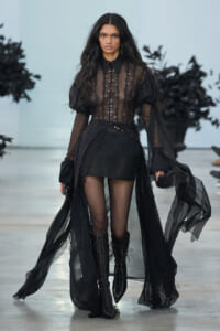 Model walking the runway in a sheer black lace blouse with a high collar, long sleeves, and a flowing lace cape over a black skirt and knee-high lace-up boots.