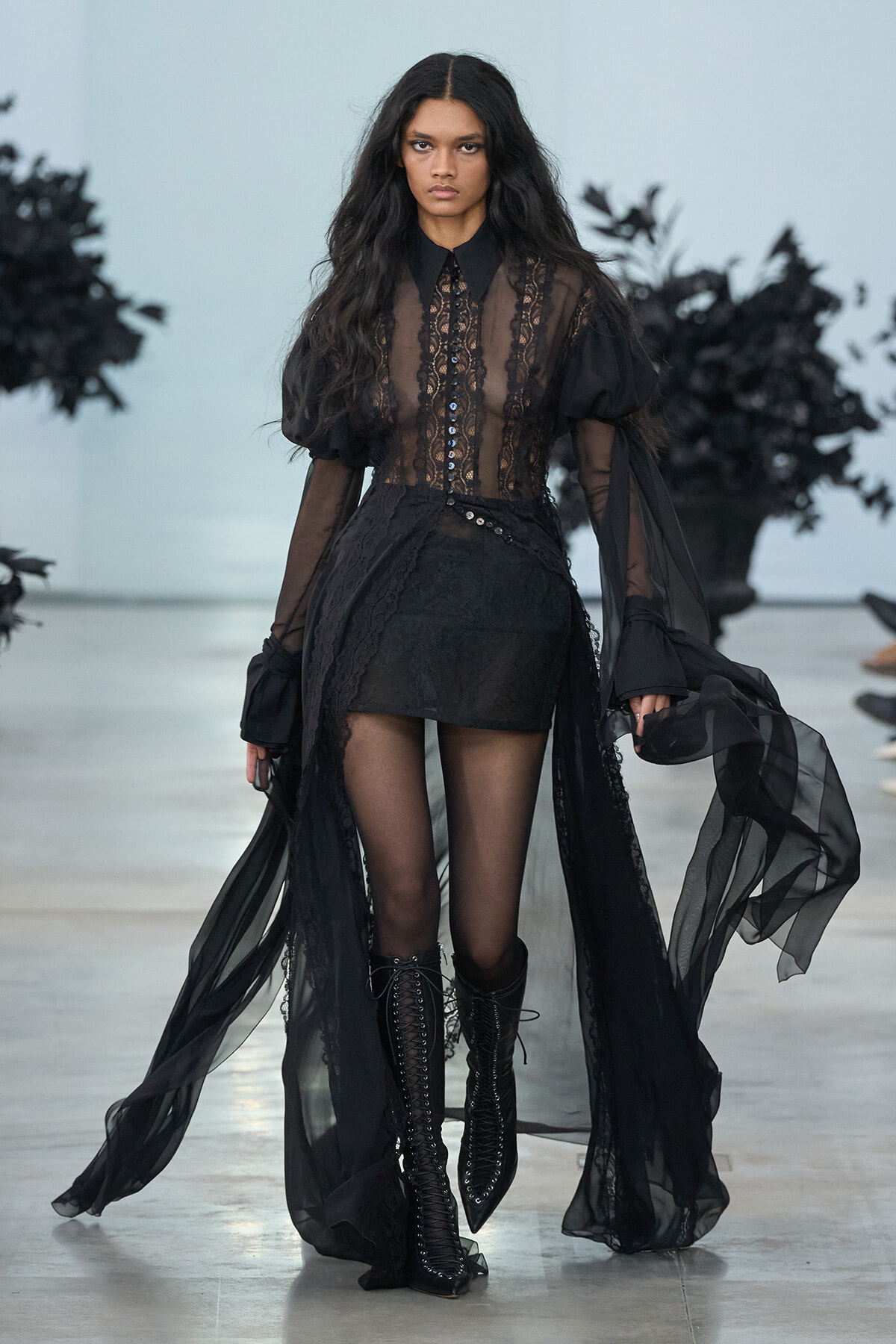 Model walking the runway in a sheer black lace blouse with a high collar, long sleeves, and a flowing lace cape over a black skirt and knee-high lace-up boots.
