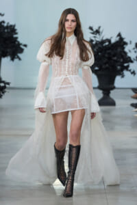 Model walking the runway in a sheer lace white dress with puff sleeves and a long train, paired with knee-high black lace-up boots