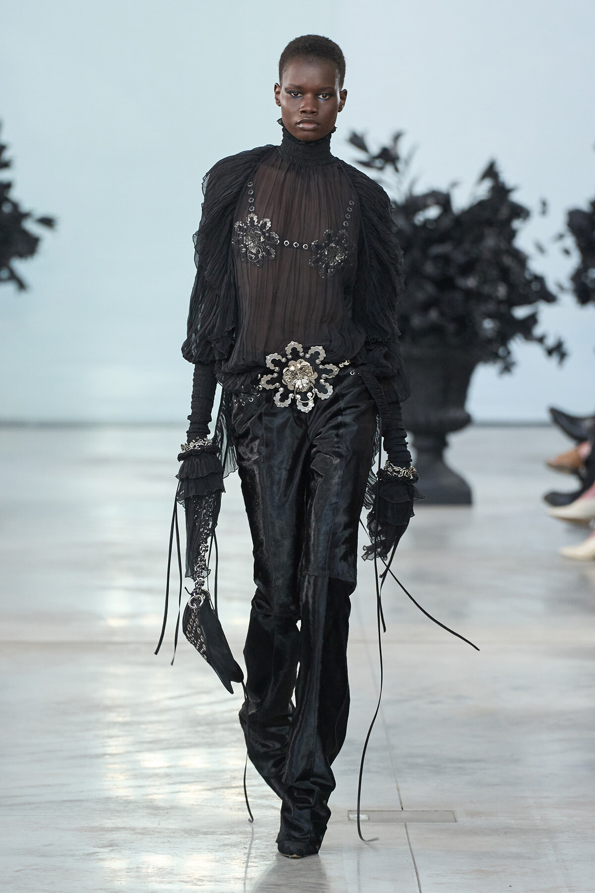 Model walks a runway in a sheer black ruffled blouse, oversized belt with silver floral embellishments, and black velvet pants with gloves.