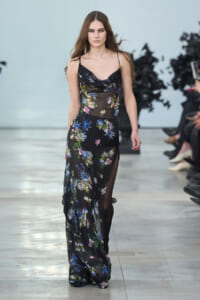 Model walking a runway in a black sheer floral gown with thin straps.