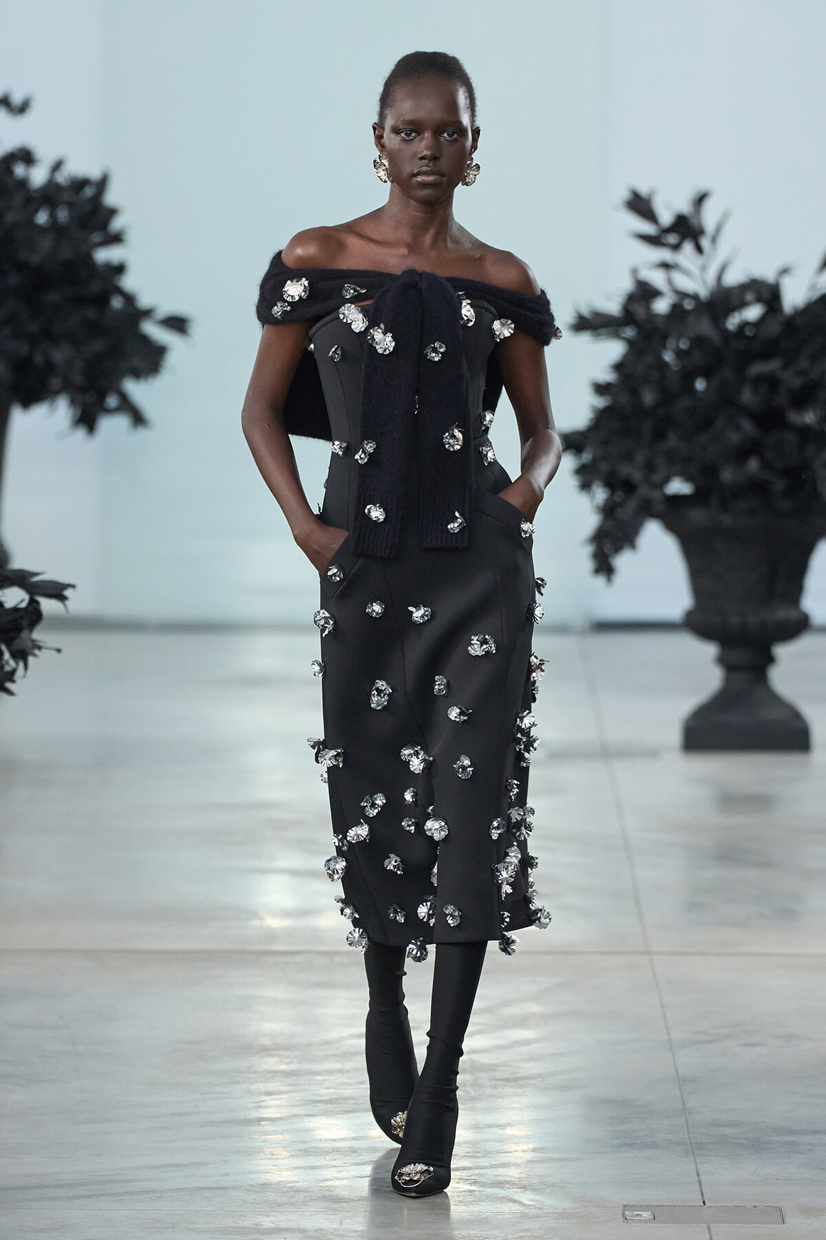 Model walks the runway in a black off-the-shoulder midi dress heavily adorned with silver crystal embellishments and a matching scarf draped over the shoulders.