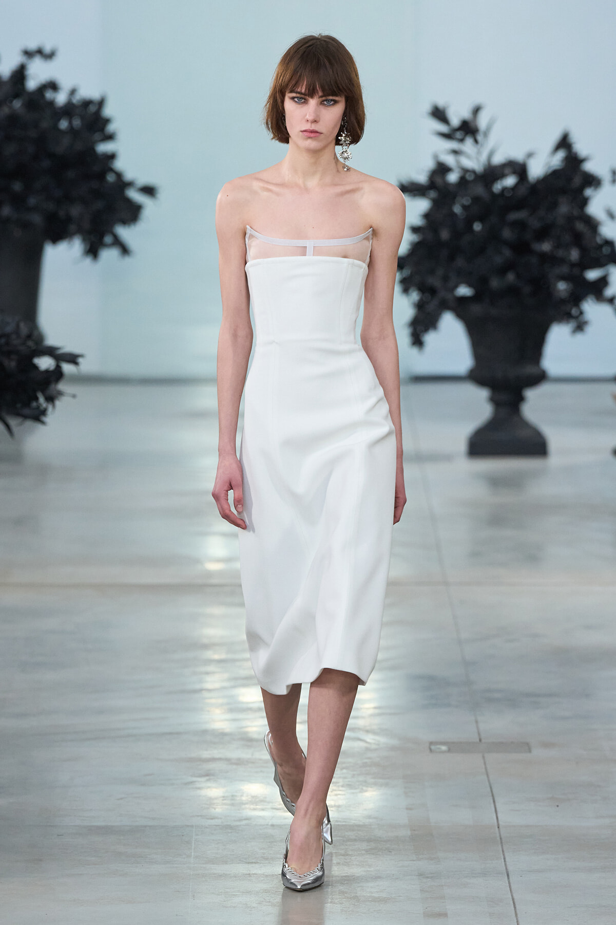Model walks a runway in a strapless white midi dress with a sheer neckline detail and silver heels.