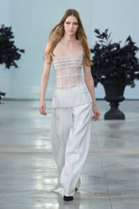 Model walks a fashion runway in a sheer white lace corset-style top with panel detailing and wide-leg white trousers.