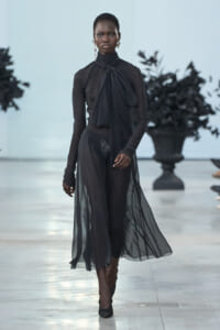 Model walking a sheer black gown with a large neck-tie knot on a runway.